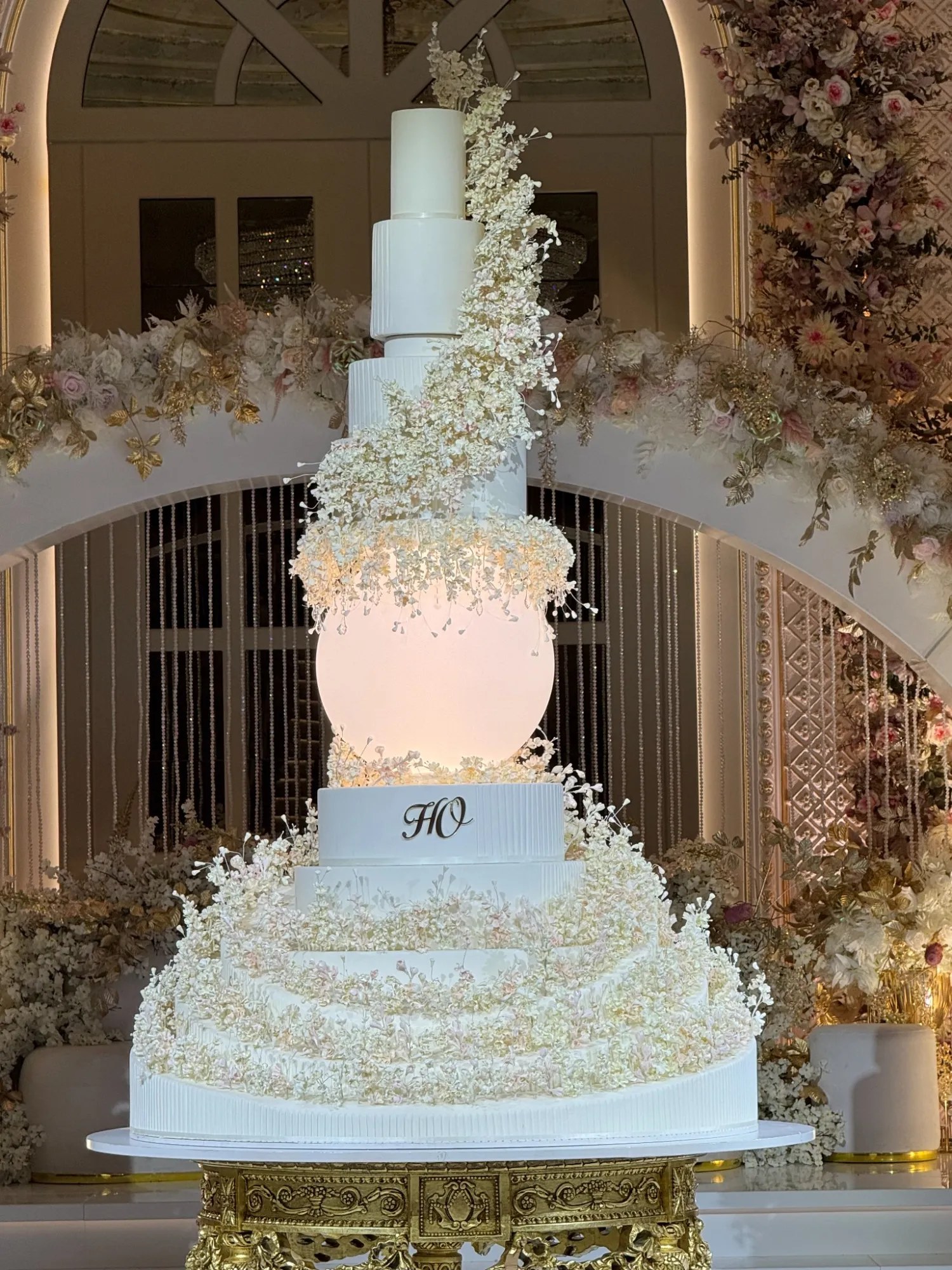 A statement wedding cake with an illuminated sphere and cascading ivory sugar flowers before a floral arch